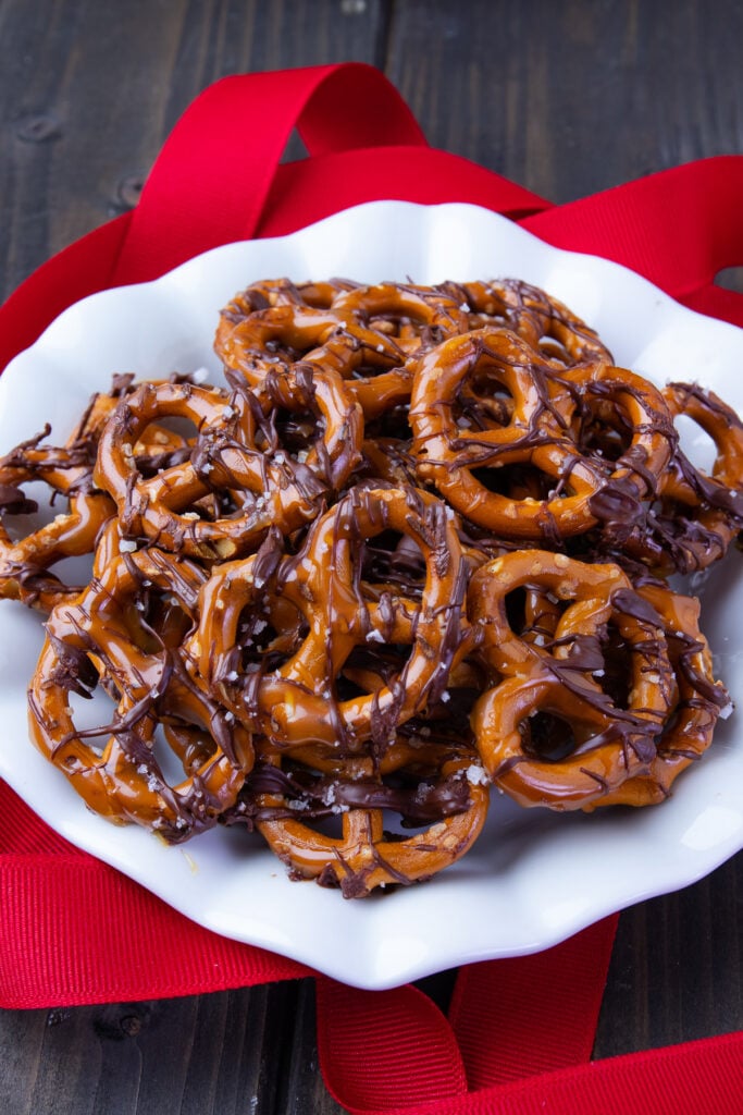 Pretzels drizzled with buttery caramel sauce and rich, dark chocolate with a pinch of flakey sea salt on a white plate with red ribbon decorations.