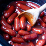 Little smokies crock pot recipe with glossy red sausages in dark slow cooker, wooden spoon stirring sauce