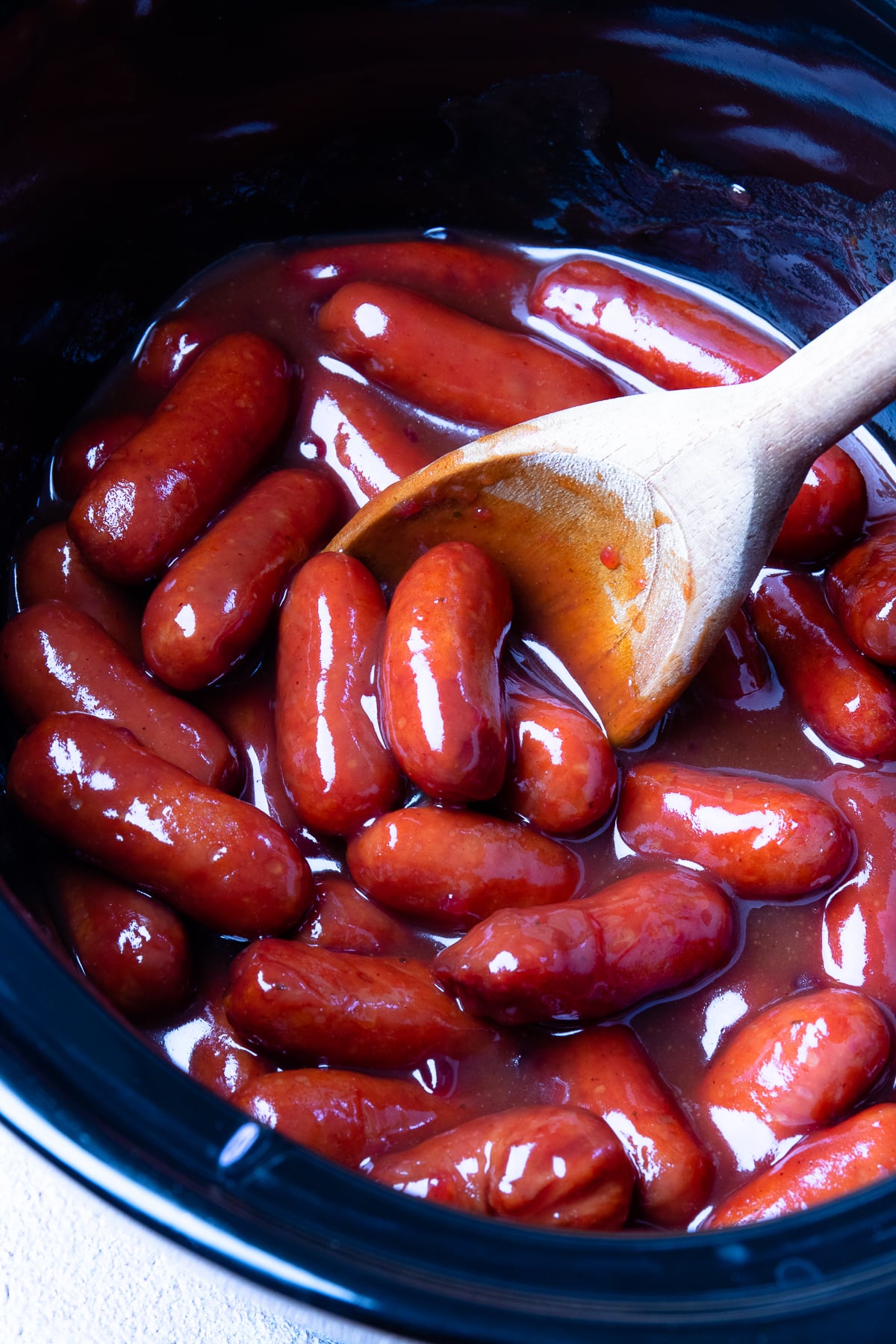 Little smokies crock pot recipe with glossy red sausages in dark slow cooker, wooden spoon stirring sauce
