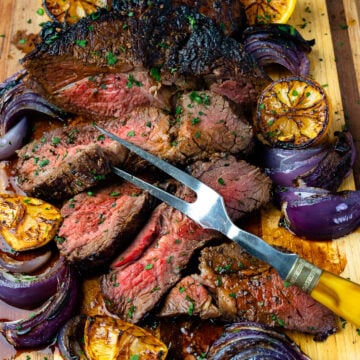 Marinated London Broil with lemons and onions with a serving fork on a wooden board.