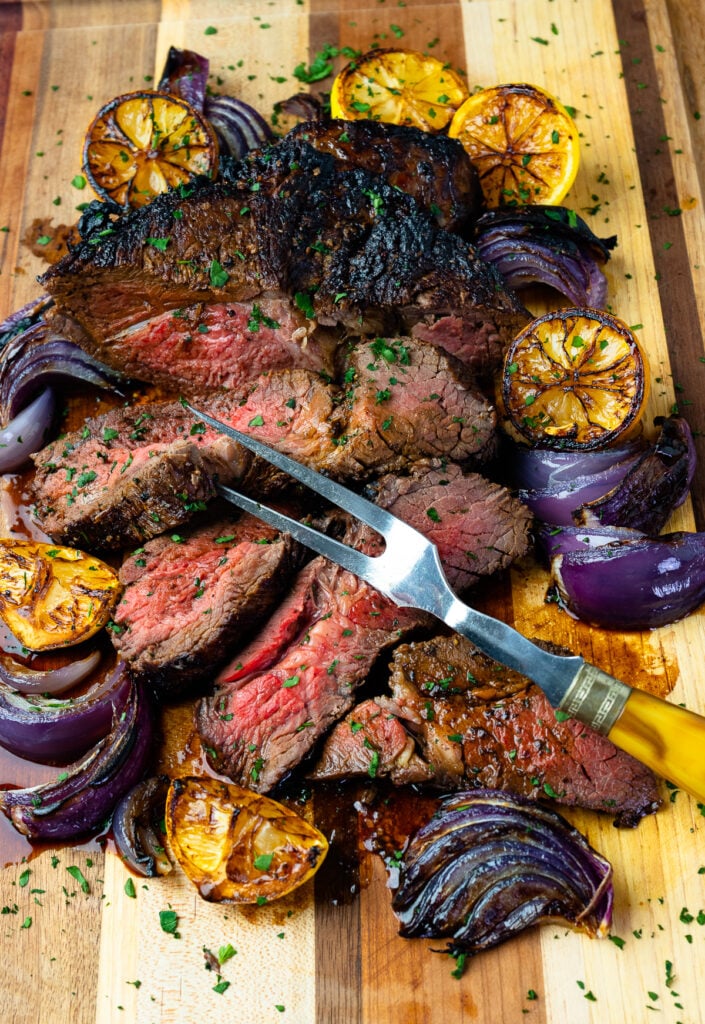 Marinated London Broil with lemons and onions with a serving fork on a wooden board.