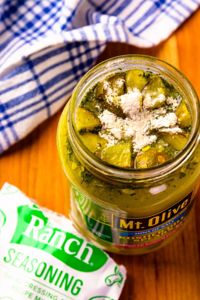 Two jars of Mt. Olive pickles on a wooden surface surrounded by Hidden Valley Ranch seasoning packets, with some seasoning spilled nearby and a blue-checked towel in the corner.