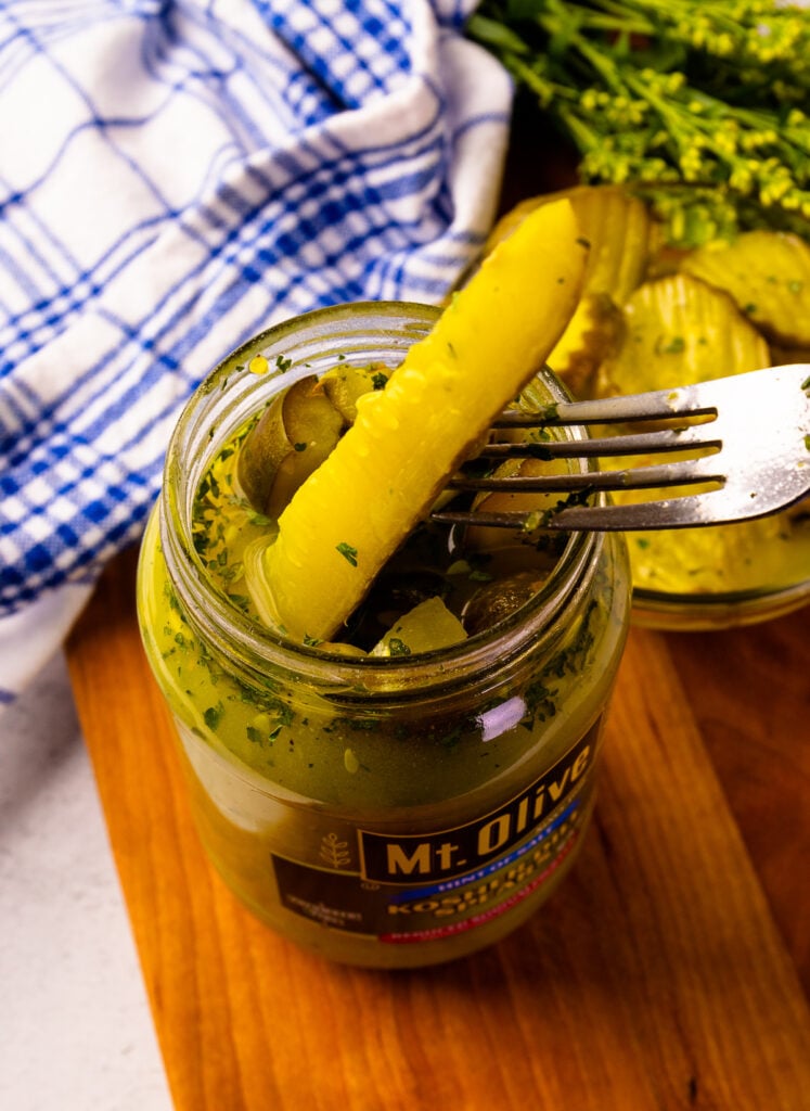 A glass bowl of sliced seasoned pickles next to an open jar of Mt. Olive pickles and a few pickle spears resting on a cutting board, garnished with herbs and a fork nearby.