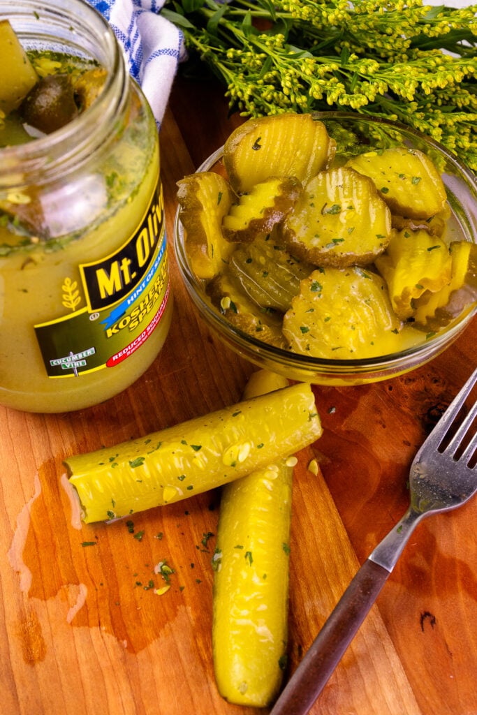 A fork lifting a dill pickle spear from a jar of Mt. Olive kosher dill pickles, seasoned with herbs. A blue and white kitchen towel and fresh herbs are in the background.