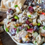 Cranberry pecan chicken salad in serving bowl with spoon, topped with diced apples, green onions, and toasted pecans on dark wood table