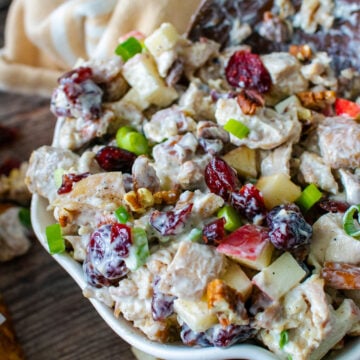 Cranberry pecan chicken salad in serving bowl with spoon, topped with diced apples, green onions, and toasted pecans on dark wood table