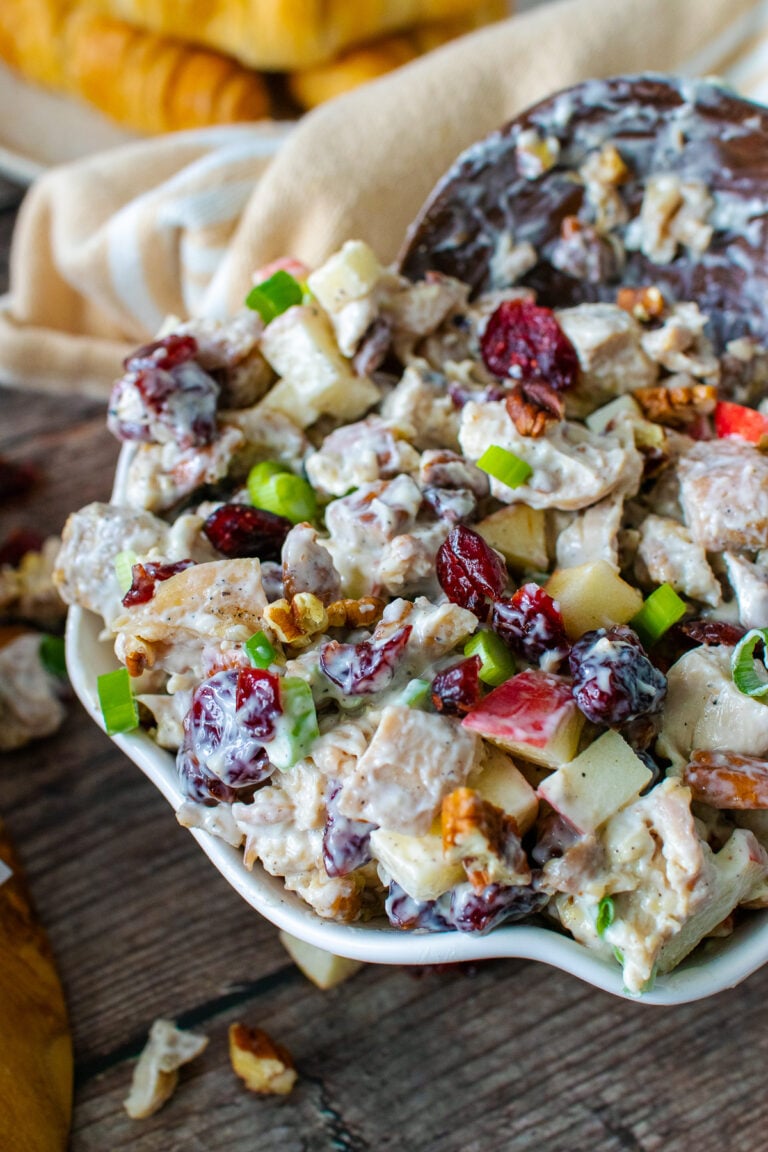 Cranberry pecan chicken salad in serving bowl with spoon, topped with diced apples, green onions, and toasted pecans on dark wood table