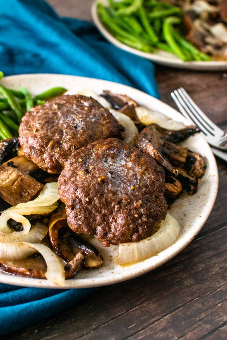 2 hamburger steak patties served with cooked onions and mushrooms and green beans on a plate.