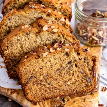 Banana bread sliced board with pecan topping on wooden serving board, jar of pecans visible beside loaf