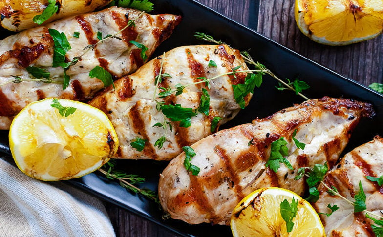 Grilled lemon chicken horz1 served on black platter with charred lemon halves and fresh parsley garnish on wooden table