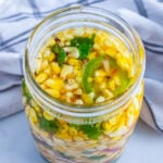 Pickled corn overhead in a glass jar with jalapeños, cilantro, garlic, and spices in oil brine on striped cloth