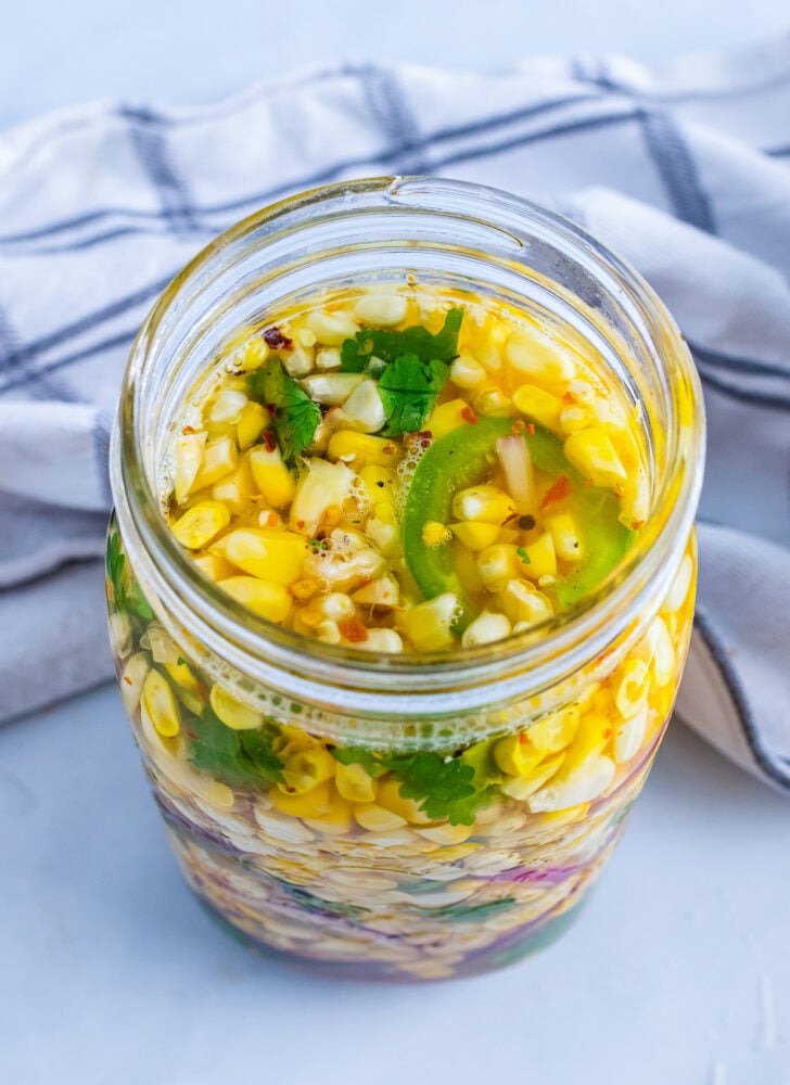 Pickled corn overhead in a glass jar with jalapeños, cilantro, garlic, and spices in oil brine on striped cloth