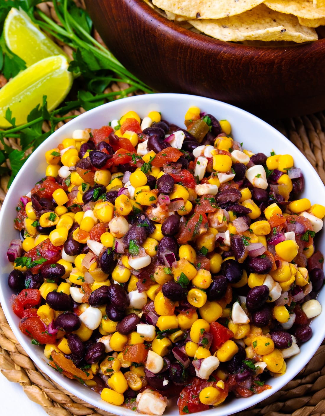 Black Bean and Corn Salsa Recipe served in white bowl with lime, cilantro, tortilla chips, and wooden serving bowl nearby