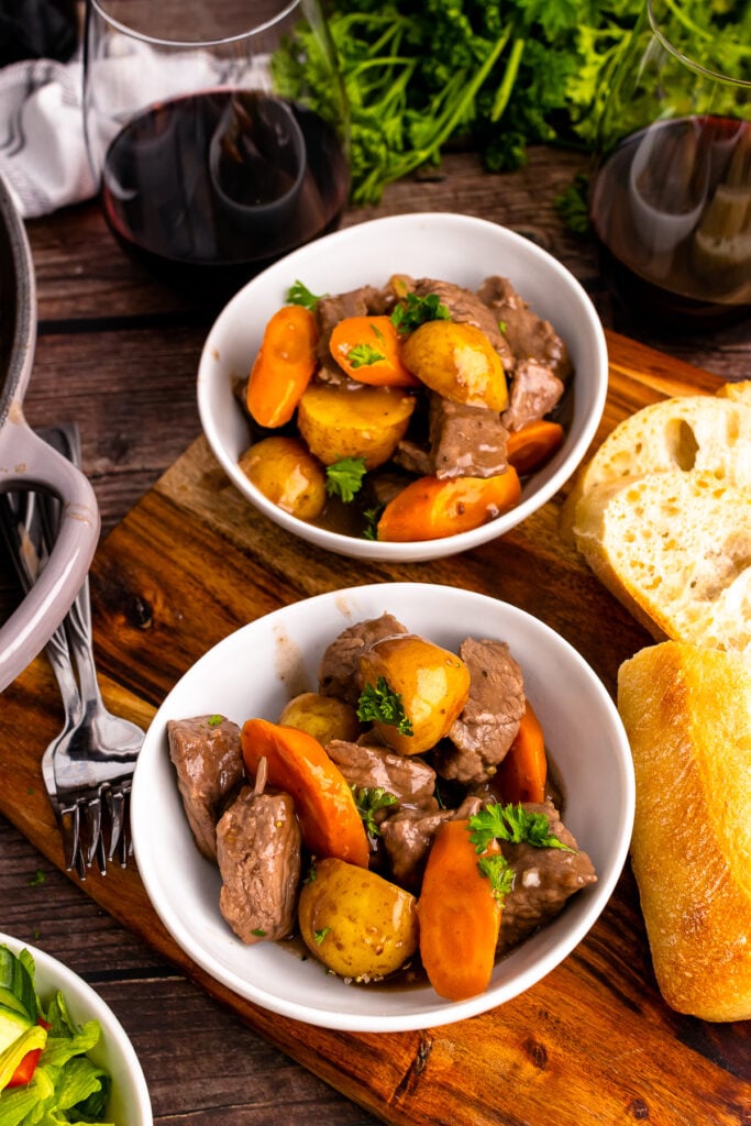 Two white bowls full of skillet beef stew served with bread and red wine.