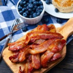 Crispy airfryer bacon strips on wooden board with fresh blueberries in white bowl, pancakes, and fork on blue checkered cloth