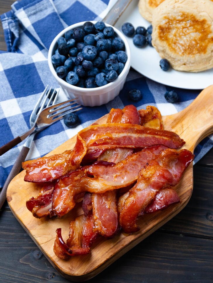 Crispy airfryer bacon strips on wooden board with fresh blueberries in white bowl, pancakes, and fork on blue checkered cloth