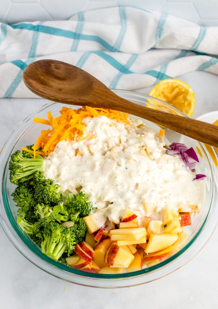 Broccoli Apple Salad ingredients in a bowl with salad dressing and a wooden spoon.