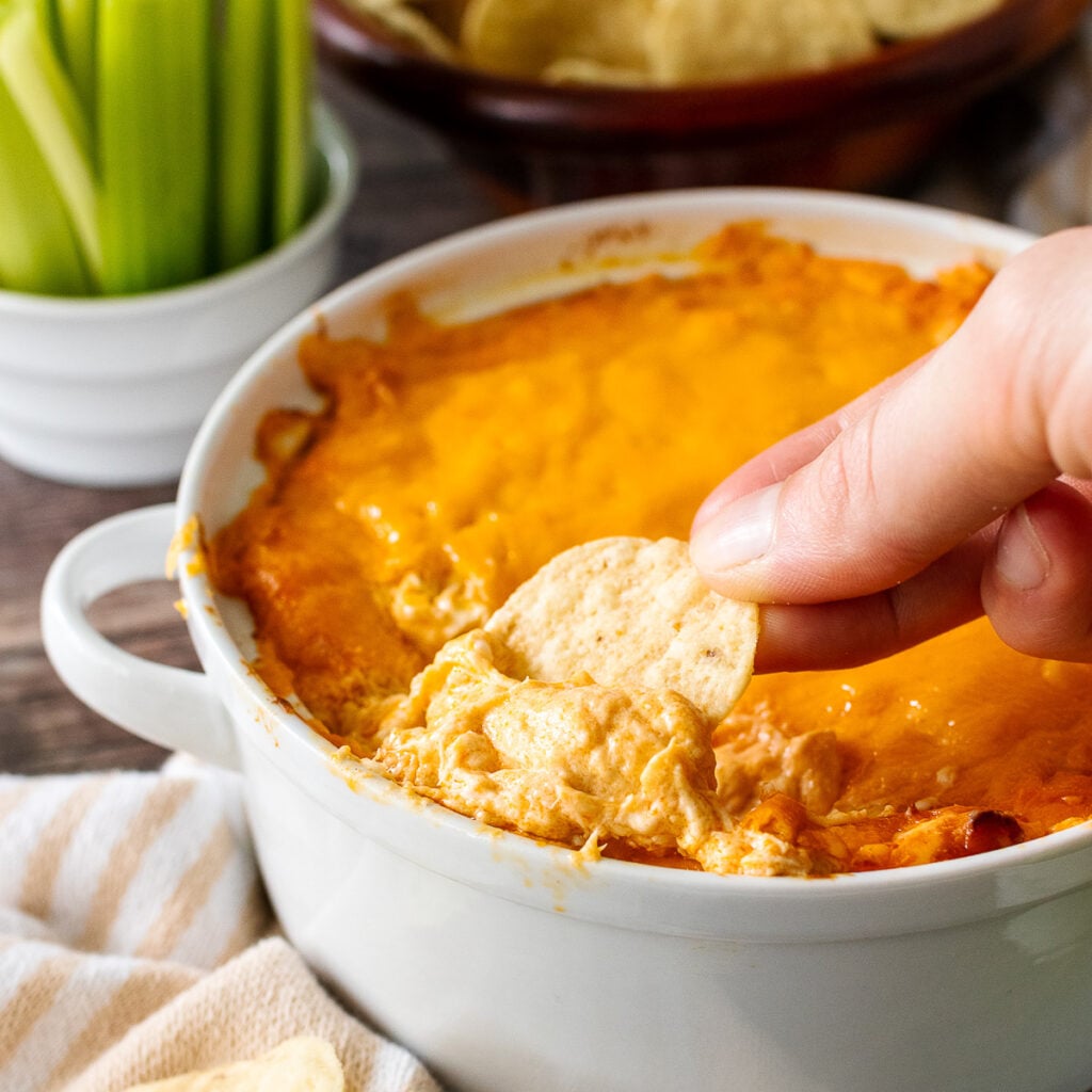Hand dipping cracker into creamy buffalo chicken dip 1200 in white bowl with fresh green onions and bread in background