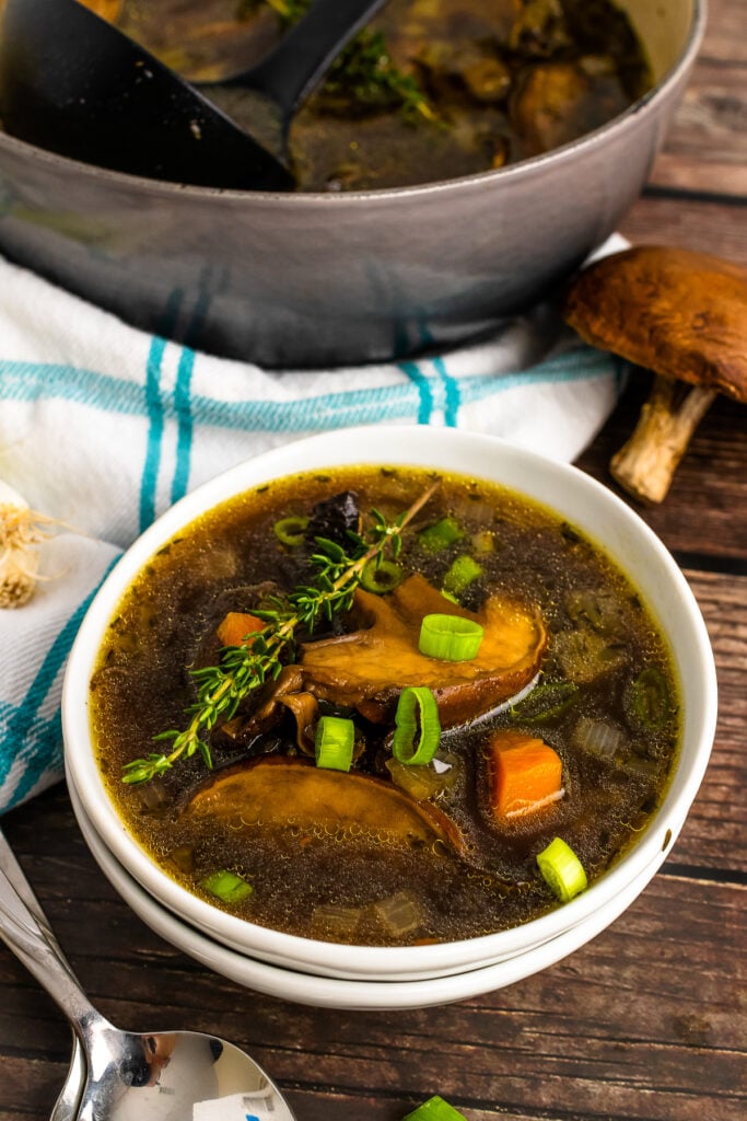 Delicious, healthy mushroom soup served in a white bowl with the stock pot sitting behind it. Topped with fresh thyme sprigs and green onions, this soup is ready to be served.