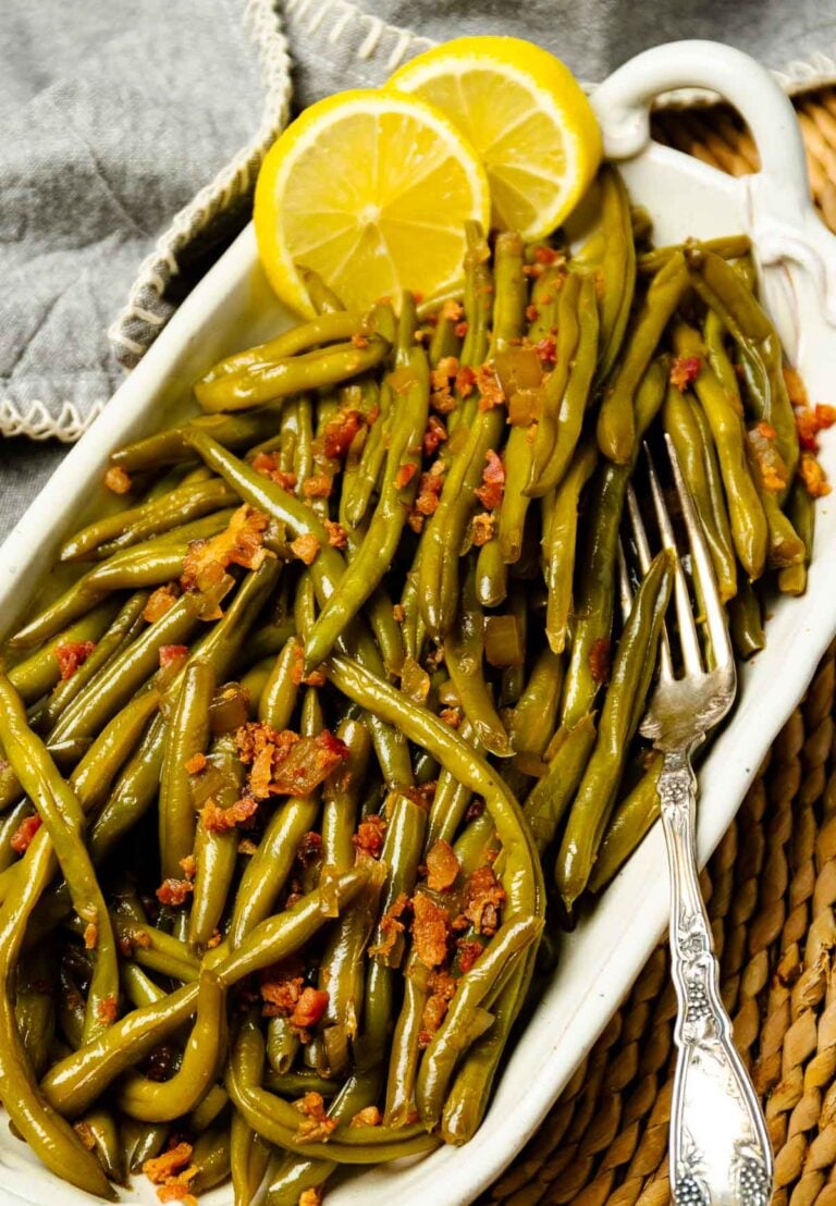 Crock Pot Maple Bacon Green Beans in a white serving dish with lemon slices and crispy bacon bits on top, with a fork beside it