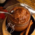 Homemade apple butter in glass jar with spoon, cinnamon sticks, and autumn ingredients on wooden board for apple butter 06
