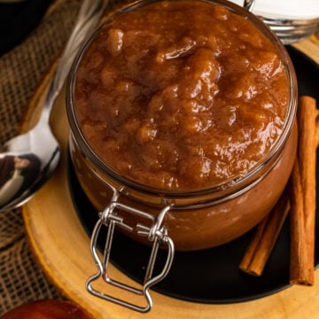 Homemade apple butter 14 in a glass jar with hinged lid on wooden board, surrounded by fresh apples and cinnamon sticks