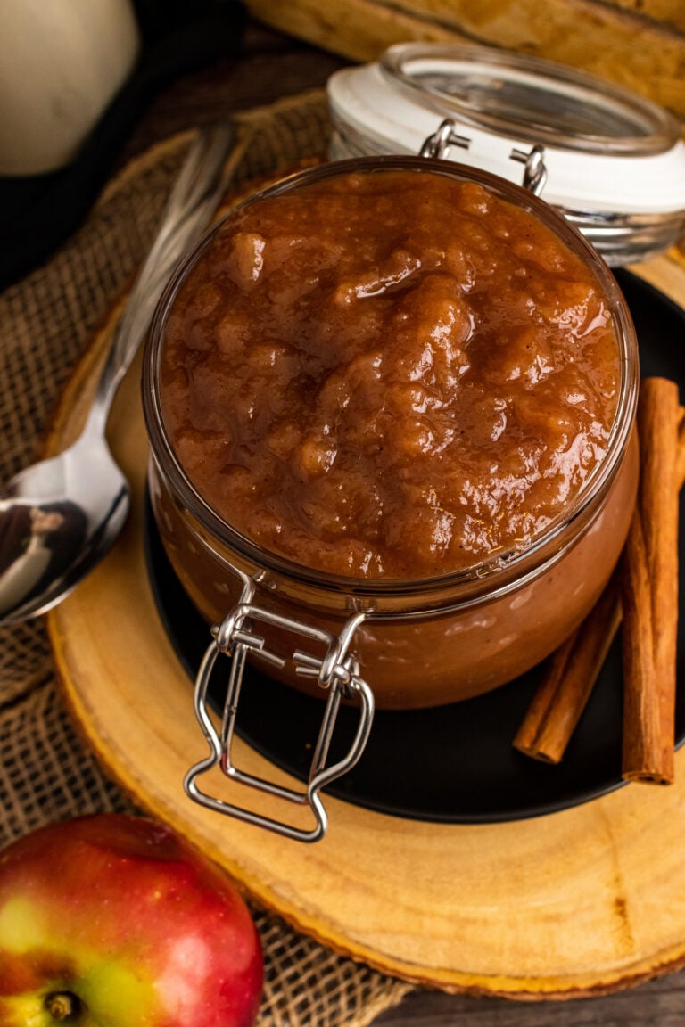 Homemade apple butter 14 in a glass jar with hinged lid on wooden board, surrounded by fresh apples and cinnamon sticks