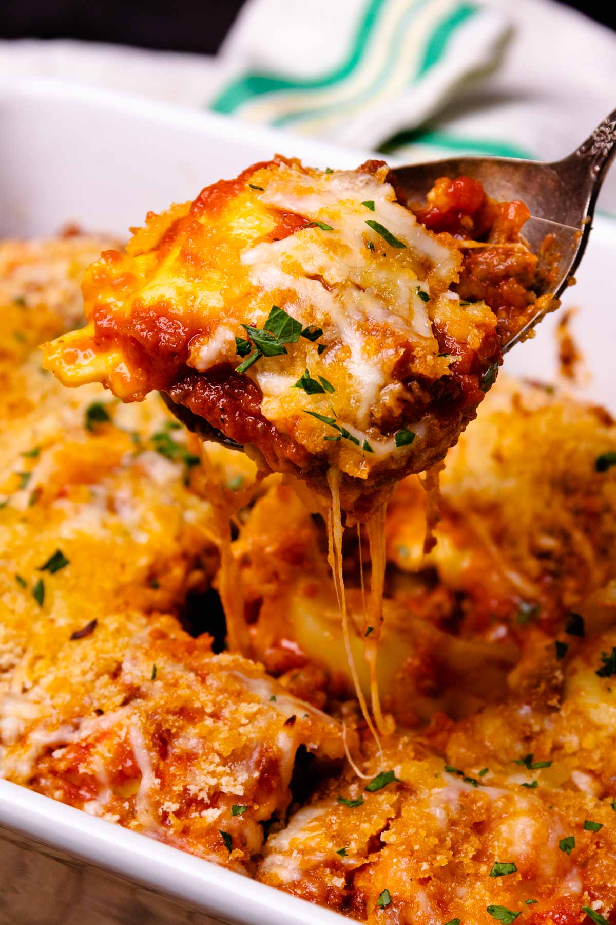 Cheesy Italian Sausage Ravioli Bake on white plate with melted cheese stretching from spoon, garnished with fresh chives