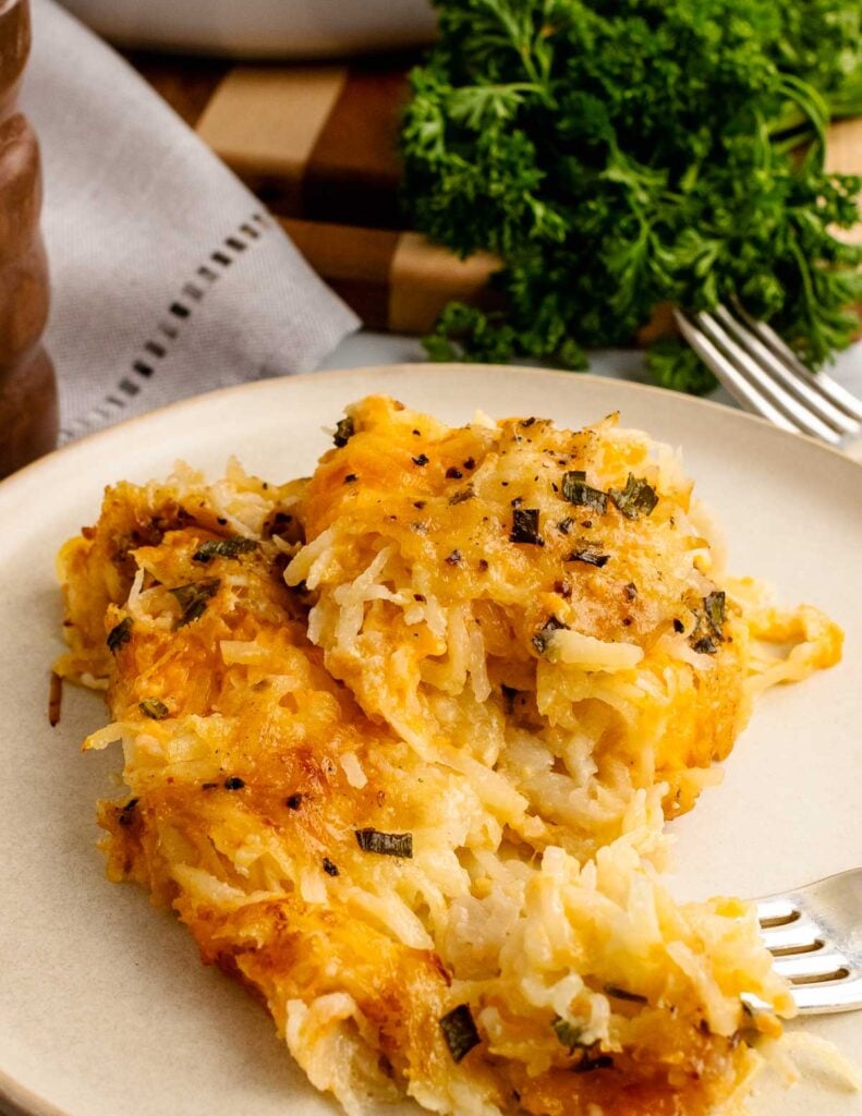 Close-up of cheesy hashbrown casserole served on a plate, topped with melted cheese and herbs, with a fork on the side