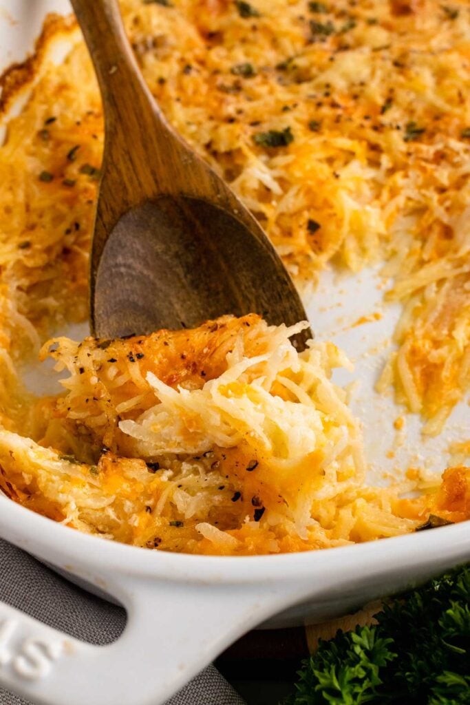 Wooden spoon scooping cheesy hashbrown casserole from a white baking dish, showing golden melted cheese and shredded potatoes.