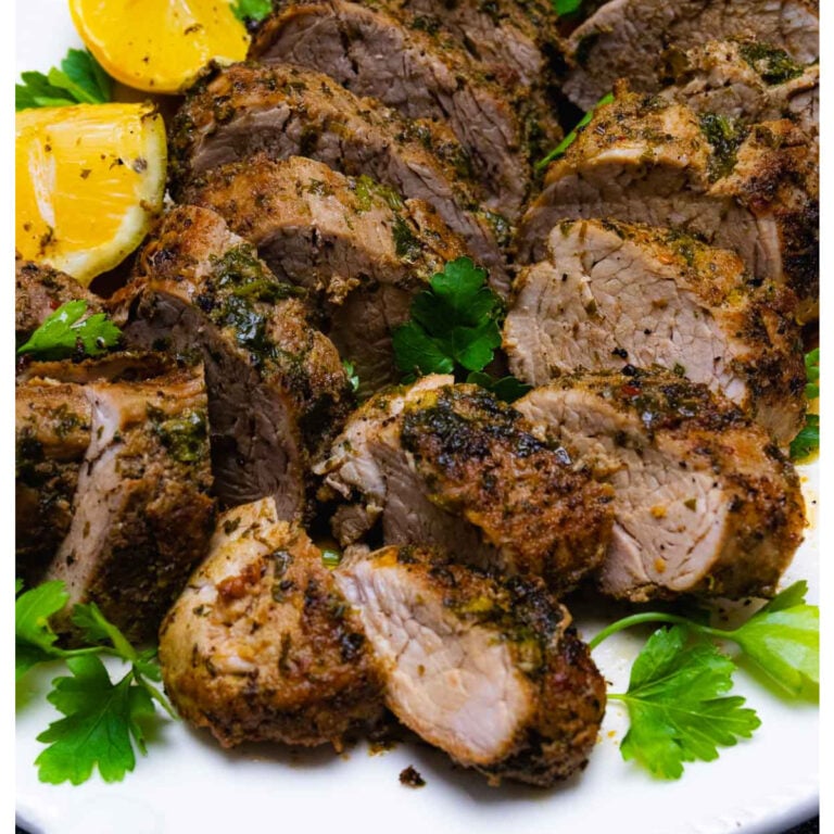 Sliced Roasted Pork Tenderloin with herb crust garnished with fresh parsley and lemon wedges on white plate
