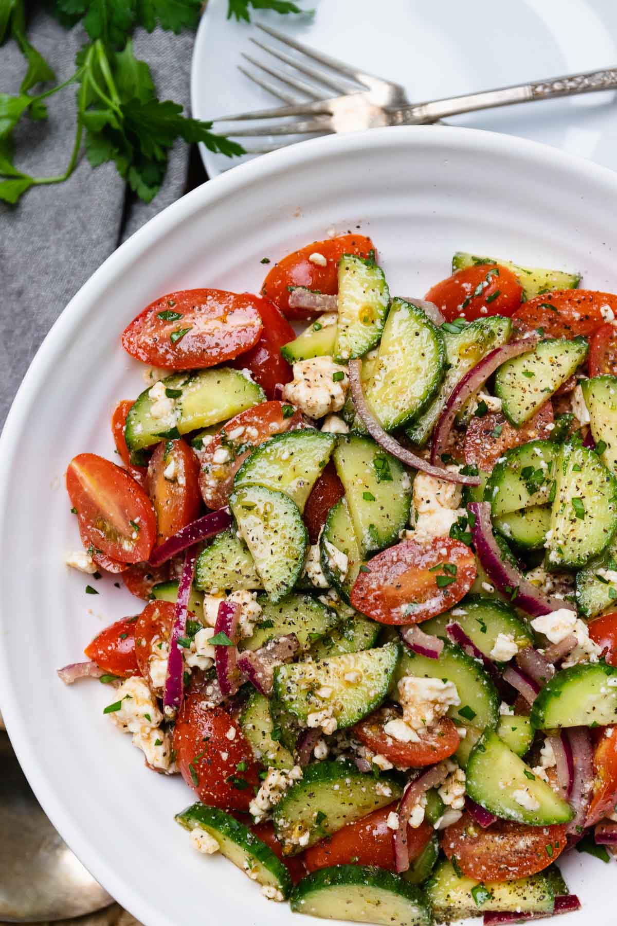 White bowl with cucumbers and tomatoes tossed in Danos homemade spicy dressing in a white bowl and forks for serving.