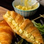 Egg salad 11 croissant sandwich on wooden plate with mixed greens, fresh greens, and bowl of egg salad behind it