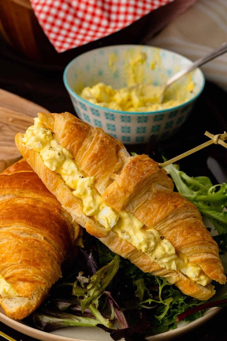 Egg salad 11 croissant sandwich on wooden plate with mixed greens, fresh greens, and bowl of egg salad behind it