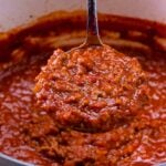 Spoon full of rich red spaghetti sauce being lifted from white pot, showing chunky meat sauce texture