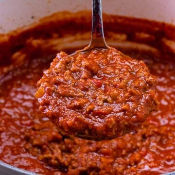 Spoon full of rich red spaghetti sauce being lifted from white pot, showing chunky meat sauce texture