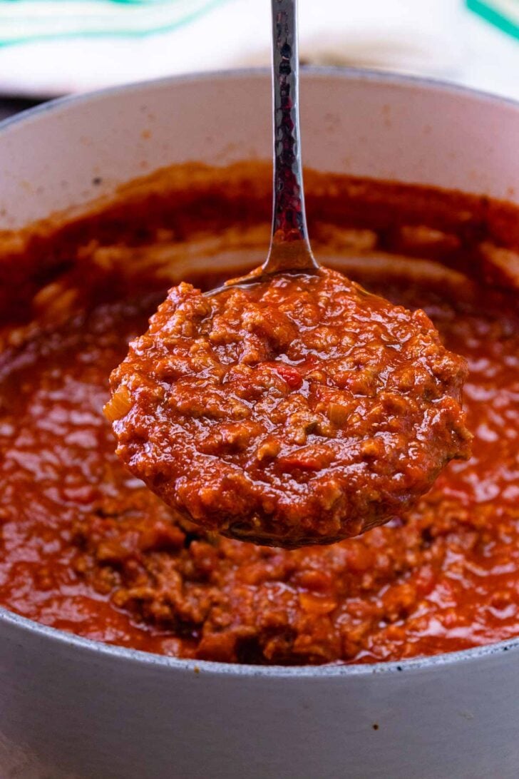 Spoon full of rich red spaghetti sauce being lifted from white pot, showing chunky meat sauce texture