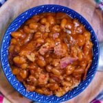 Easy Crock Pot Baked Beans in blue ceramic bowl with bacon pieces, held in hands over colorful cloth background