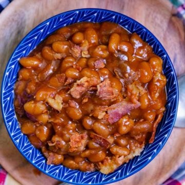 Easy Crock Pot Baked Beans in blue ceramic bowl with bacon pieces, held in hands over colorful cloth background