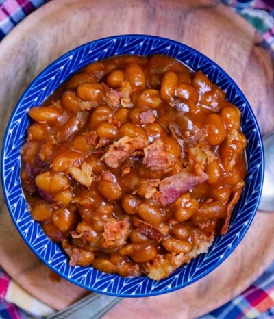 Easy Crock Pot Baked Beans in blue ceramic bowl with bacon pieces, held in hands over colorful cloth background