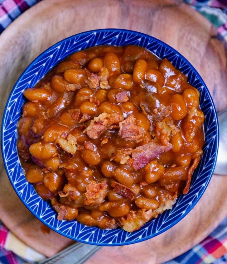 Easy Crock Pot Baked Beans in blue ceramic bowl with bacon pieces, held in hands over colorful cloth background