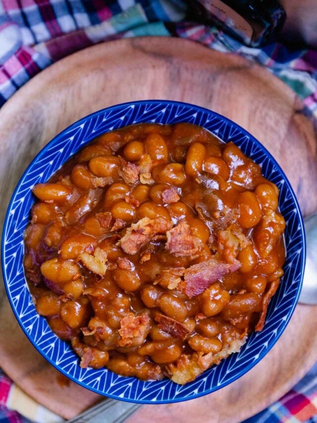 Cropped close-up of baked beans in blue-rimmed bowl topped with crispy bacon pieces on wooden charger with checkered cloth