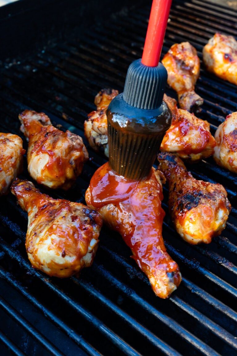Glazing chicken pieces on grill grates with Homemade BBQ Sauce (Easy And Delicious) using a red-handled brush, charred and caramelized