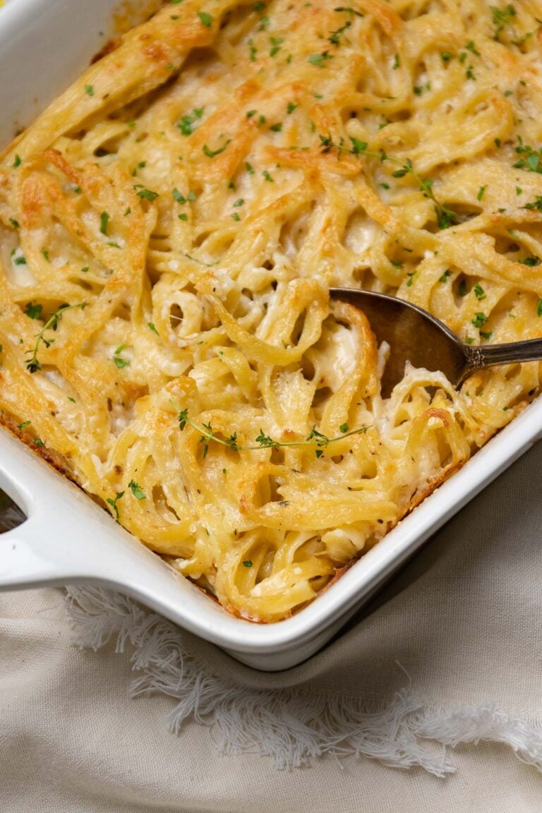 Creamy Baked Fettuccine With Cheese in white baking dish, garnished with fresh parsley and black pepper, with serving spoon