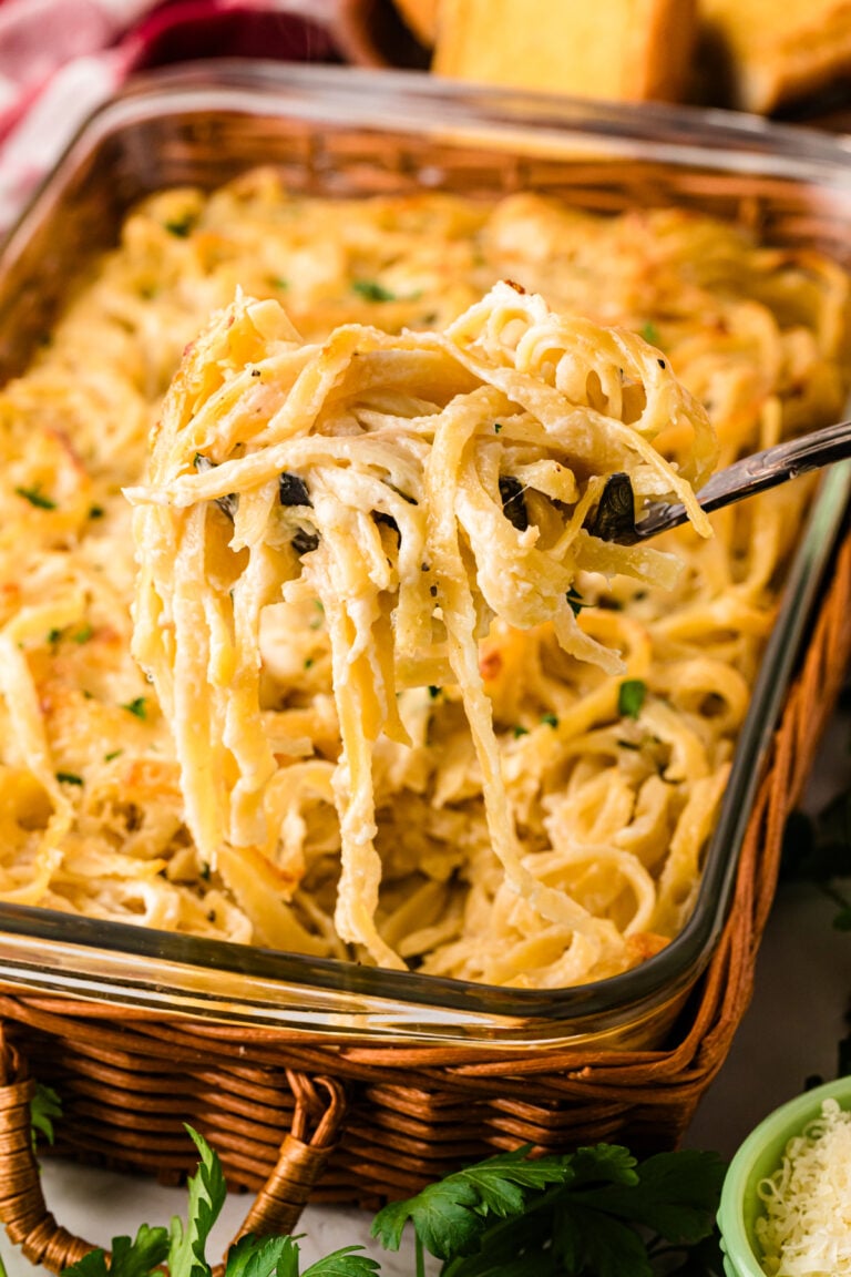 Creamy Baked Fettuccine With Cheese being lifted on a fork from a glass baking dish lined with a woven basket holder, garnished with fresh p