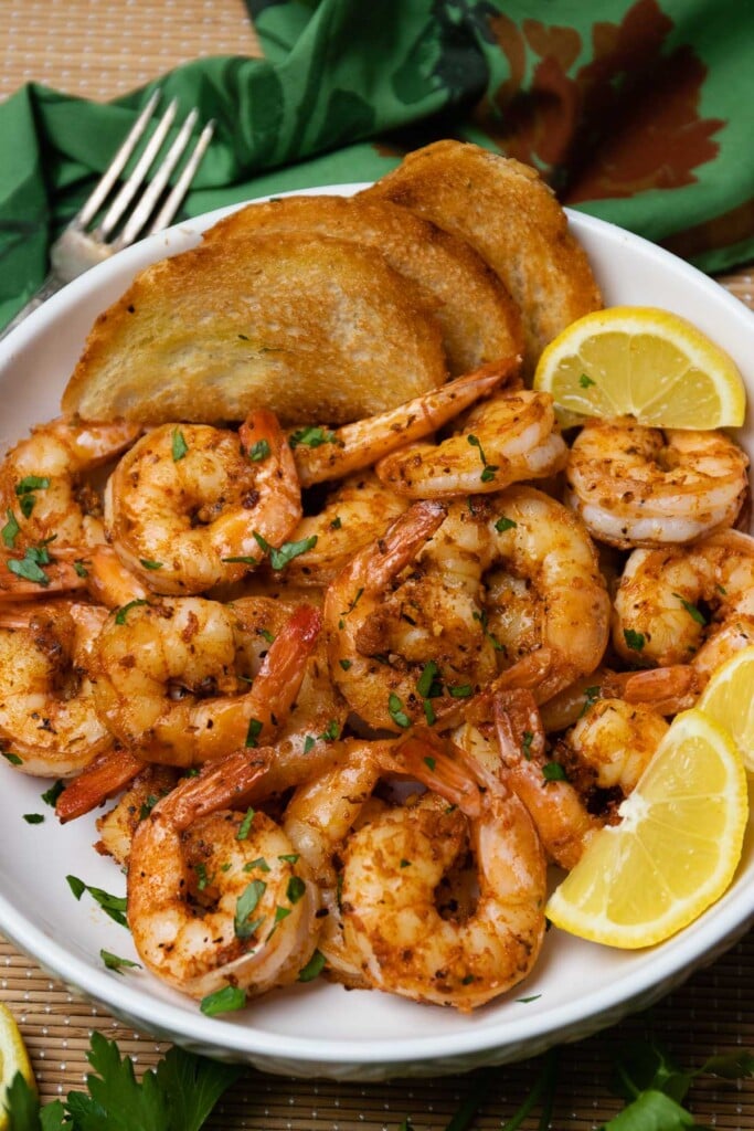 Beautiful white bowl filled with cooked cajun garlic shrimp served with toast and lemons then garnished with fresh parsley as an appetizer or main dish.