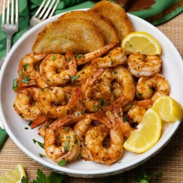 Beautiful white bowl filled with cooked cajun garlic shrimp served with toast and lemons then garnished with fresh parsley.