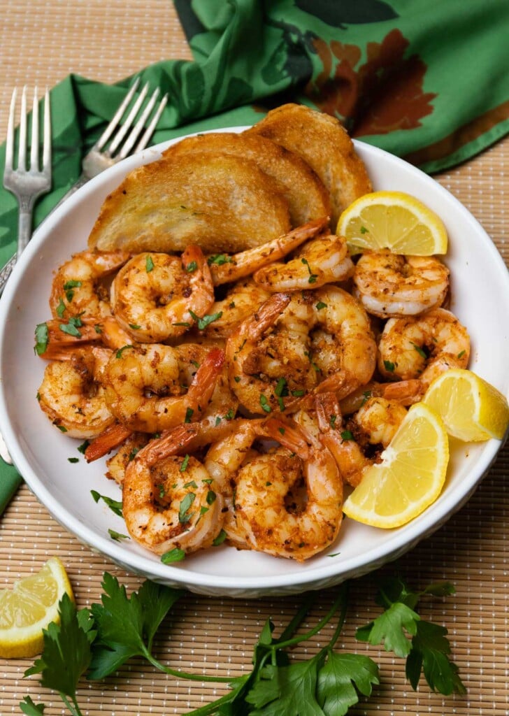 Beautiful white bowl filled with cooked cajun garlic shrimp served with toast and lemons then garnished with fresh parsley.