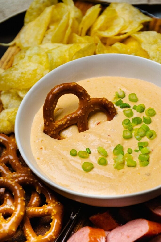 Creamy cheese dip with pretzel and green onion garnish surrounded by potato chips, pretzels, and sausage for Easy Slow Cooker Dinners for Bu