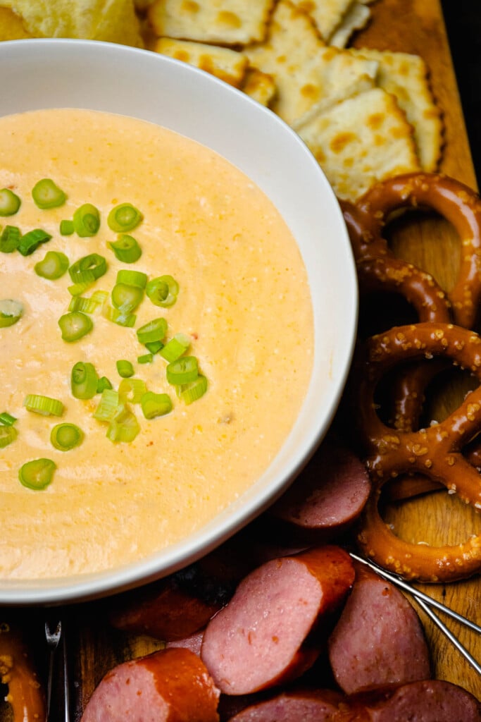 Large white bowl with Beer Cheese Dip topped with green onions served with sausage and pretzels.